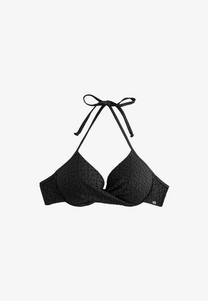 Kvinde med langt mørkt hår iført sort bikini står på en sandstrand med blåt hav og grønne bakker i baggrunden.