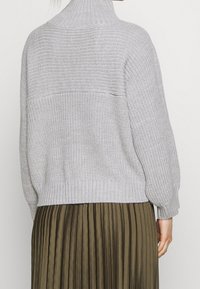 Pull gris clair en tricot avec un col montant et une ceinture côtelée, associé à une jupe plissée vert olive foncé.