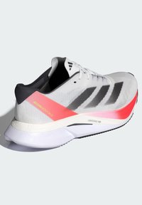 Zapatilla deportiva blanca con rayas negras y acentos en rosa, que presenta una parte superior de malla texturizada, un collar acolchado y una suela ligera.