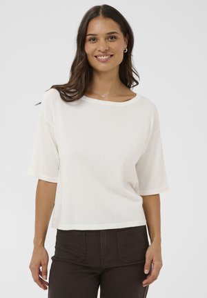 Kvinde med langt brunt hår iført hvid kortærmet top, mørkebukser, sølvhalskæde og creoler, stående foran en ensfarvet hvid baggrund.