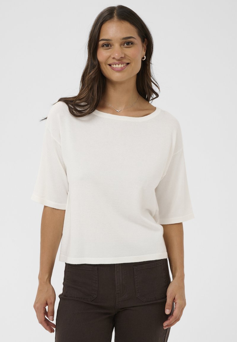 Femme aux longs cheveux bruns portant un haut blanc à manches courtes, un pantalon foncé, un collier en argent et des boucles d'oreilles créoles, debout devant un fond blanc uni.