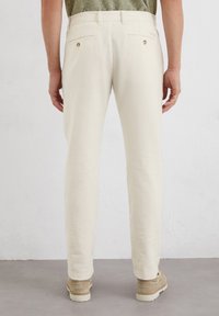 Beige linen pants with a straight-leg design, featuring rear pockets with gold-tone buttons and a flat waistband, paired with tan shoes.