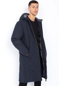 Mann in langem, navyblauen Wintermantel mit Kapuze, schwarzem Hemd und Hose, posiert mit den Händen in den Taschen vor einem weißen Hintergrund.