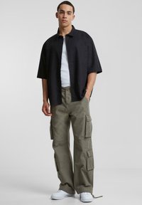 Cămașă neagră cu mâneci scurte, cu nasturi, peste un top alb, asociată cu pantaloni cargo de culoare verde oliv, care au buzunare laterale, și adidași albi.