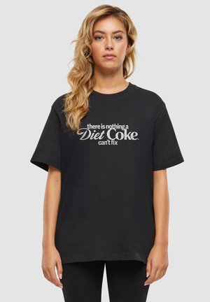 Femme aux longs cheveux blonds portant un t-shirt noir avec un texte blanc disant "there is nothing a Diet Coke can't fix" sur un fond gris uni.