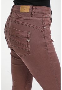 Jeans de mezclilla marrones con un diseño ajustado, que cuentan con una etiqueta de cuero marrón en la cintura, costuras decorativas y dos bolsillos traseros.