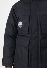 Giubbotto imbottito nero con multiple tasche frontali, chiusura a bottoni e patch circolare con logo sul petto. Presenta un tessuto testurizzato.