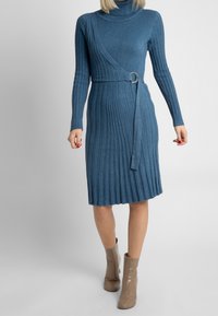 Robe en tricot côtelé bleu avec un col haut, un design enveloppant, une ceinture latérale avec une boucle circulaire et un ourlet évasé. Assortie avec des bottines beiges.