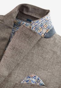 Brauner strukturierter Blazer mit umgeschlagenem Kragen und Taschentuch, beide mit komplexen Blumenmustern in Blau, Orange und Beige.