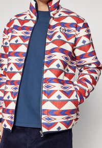 Fleece zip-up jack met een geometrisch patroon in rood, blauw en wit. Hoge kraag, zijzakken, contrasterende blauwe binnenkant.