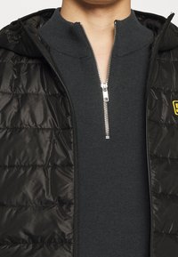 Černá péřová vesta s předním zipem, nošená přes tmavě šedý svetr s vysokým límcem. Má prošívanou strukturu a stříbrný zip.