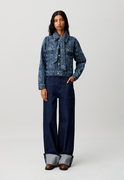 Donna che indossa una giacca di jeans con motivo paisley e jeans scuri a gamba larga risvoltati sopra scarpe marroni, in piedi con la mano in tasca.
