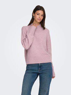 Kvinde iført en lyse-lilla strikket sweater med perleudsmykninger på krave og manchetter, kombineret med blå jeans, stående mod en hvid baggrund.