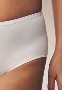 Slip en coton blanc avec une texture côtelée douce et une ceinture portant le nom de la marque "NEXT." Vue rapprochée mettant en valeur l'ajustement et le design.