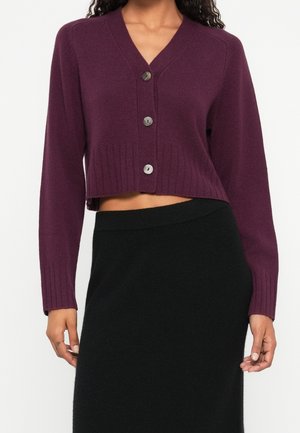 Femme portant un cardigan court violet boutonné avec poignets et ourlet côtelés, associé à une jupe noire taille haute.