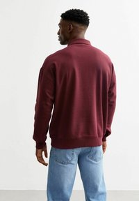 Bordeaux sweatshirt met een kraag, ribbel manchetten en boord. Draag het over lichtblauwe jeans, bekeken vanuit de achterkant tegen een effen achtergrond.