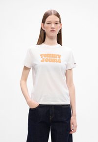 Biała bawełniana koszulka z dużym pomarańczowym logo "TOMMY JEANS" w zaokrąglonym kroju, zestawiona z ciemnoniebieskimi jeansami o wysokim stanie.