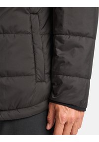 Giacca a vento scura con un design trapuntato, texture morbida e una tasca laterale. Il polsino ha un rivestimento interno nero a contrasto.