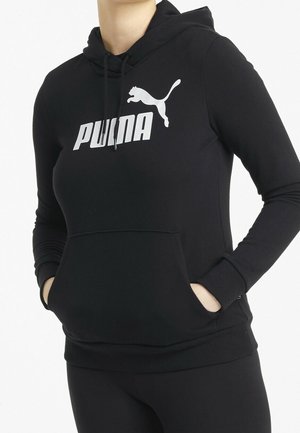 Person trägt einen schwarzen Puma-Hoodie mit den Händen in der vorderen Tasche, mit weißem Puma-Logo und Text auf der Brust.