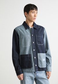 Jeansjakke med fargeblokker i mørk og lys blå med store frontlommer, hvite knapper og en firkantet merkeetikett på den nedre lommen.