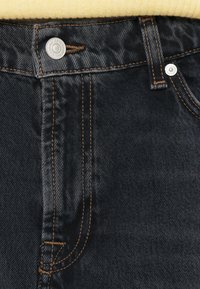 Tumedast teksast teksapüksid, millel on kontrastne oranž õmblus, hõbedane nööbi sulgemine ja puhas, klassikaline disain, millel on viis taskut.