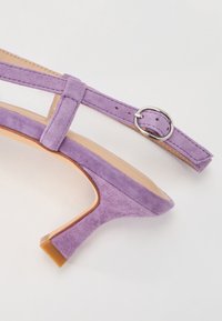 Sandale à talons hauts en suede violet avec une sangle à boucle, présentant un bout pointu et un talon fin sur une semelle beige.