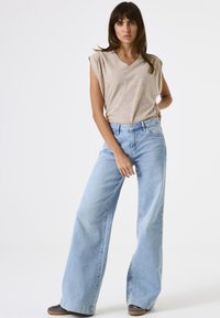 Top de manga corta beige a rayas con escote en V, combinado con jeans de pierna ancha de color azul claro. El modelo está de pie de manera casual, usando zapatos oscuros.