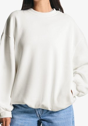 Vrouw draagt een losse witte trui met geribde manchetten en kraag, gecombineerd met lichtblauwe jeans, staand tegen een witte achtergrond.