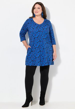Blaue Tunika mit Blumenmuster, Dreiviertelärmel, runder Ausschnitt, seitlichen Taschen, kombiniert mit schwarzen Leggings und Stiefeletten.