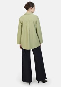 Camisa verde de manga larga con cuello y pliegue en la parte trasera, combinada con pantalones vaqueros oscuros de pierna ancha y tacones negros de punta.