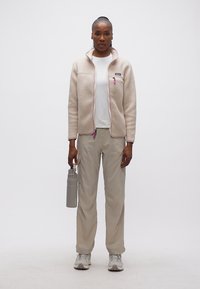 Γυναίκα με μπεζ φλις μπουφάν και ανοιχτόχρωμο παντελόνι που κρατά γκρι παγούρι νερού, στέκεται μπροστά σε απλό λευκό φόντο.