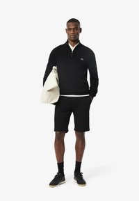 Sweater noir à zip, col blanc, associé à un short noir. Tient un sac clair, porte des chaussettes noires et des baskets.