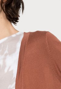 Cardigan color ruggine con una texture morbida e maniche corte, abbinato a una maglietta leggera con stampa tie-dye. Il tessuto a maglia presenta un design a costine.