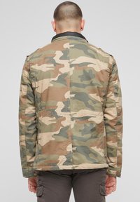 Jachetă de camuflaj în nuanțe de verde, maro și bej. Prezintă o textură căptușită și un guler negru. Buzunarele sunt vizibile pe părți.