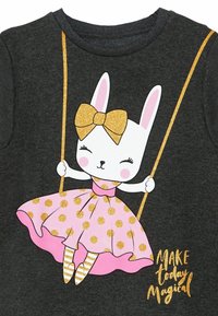 Ciemnoszara koszulka z białym królikiem w różowej sukience w kropki i złotą kokardą, huśtającym się na żółtych linach, z złotym tekstem poniżej.