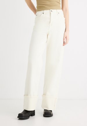 Femme portant un pantalon blanc large avec revers, un haut beige sans manches, et des bottes noires à talons, debout devant un fond blanc.