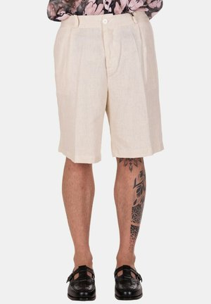Pantaloncini beige plissettati al ginocchio indossati con mocassini neri intrecciati e tatuaggio visibile sul polpaccio sinistro su sfondo uniforme.