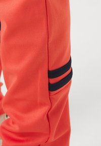 Rød atletisk jakke med en glat tekstur, der har sorte striber på ærmet. Designet er tætsiddende med en ren sømfinish.