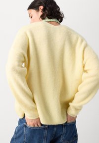 Gelb gestrickter Pullover mit einer weichen, strukturierten Oberfläche, der落ichtere Schultern und eine lockere Passform hat. Die Rückansicht zeigt den gerippten Saum und die Bündchen.