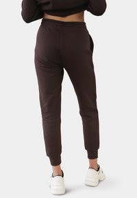 Tmavohnědé tepláky s elastickým páskem a zúženými manžetami, vyrobené z měkké tkaniny, v kombinaci s bílými sportovními botami.