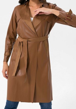 Femme portant un trench-coat marron en similicuir avec ceinture et détails de boucles aux manches, par-dessus un jean bleu.