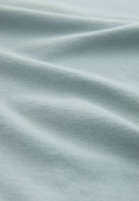 Gaiši zils kokvilnas audums ar gludu tekstūru, rādot smalkas viļņošanās un locīšanas pazīmes, kas liecina par viegla apģērba materiālu.