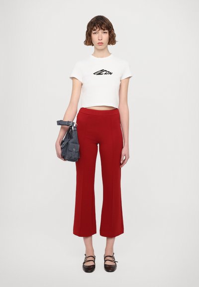 T-shirt court blanc avec un logo noir, associé à un pantalon rouge évasé et un sac à main grisé texturé. Chaussures brunes avec des lanières.