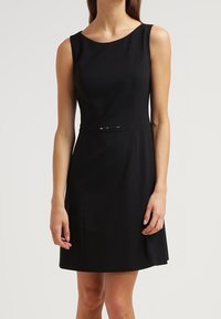 Vestido negro sin mangas con escote redondo, corsé ajustado y falda acampanada. Presenta un delgado acento plateado en la cintura. Textura de tela suave.