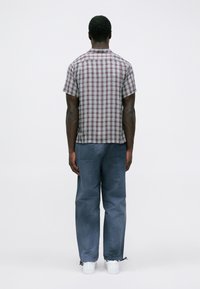 Uomo di spalle che indossa una camicia a maniche corte a quadri rossi e bianchi, pantaloni blu larghi e scarpe bianche su uno sfondo grigio uniforme.