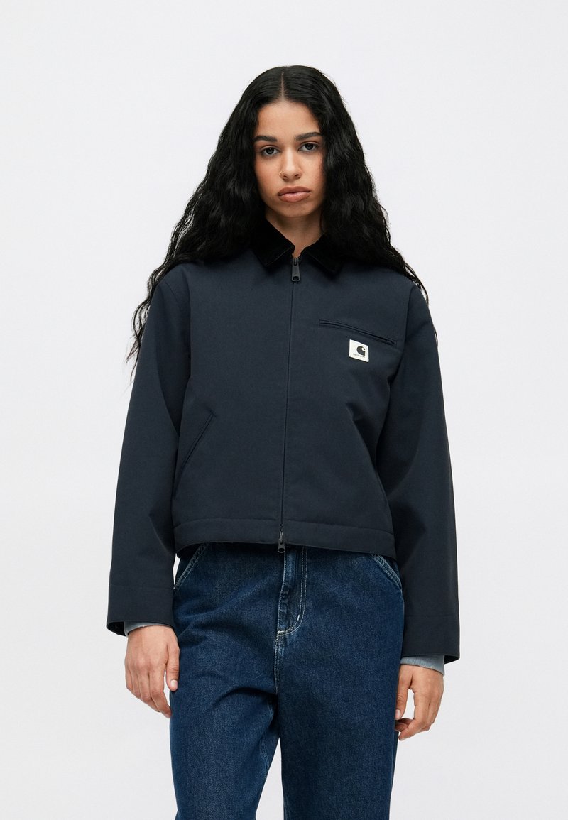 Marineblå cropped jakke med krave, lynlås lukning og frontlomme på brystet. Har logo-mærke og et glat, tekstureret materiale.