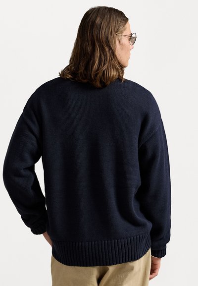 Man med axellångt brunt hår och glasögon, klädd i en marinblå stickad tröja och beige byxor, vänd bortåt mot en neutral bakgrund.