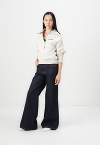 Beige zip-up sweatshirt with a high collar, paired with dark wide-leg jeans. The outfit has a relaxed fit and casual design.