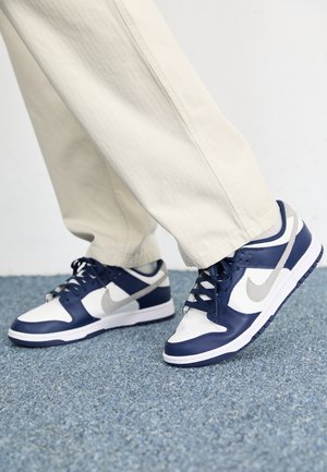 Osoba nosząca białe i granatowe buty Nike z szarym logo, stojąca na niebieskim dywanie, ubrana w beżowe spodnie.
