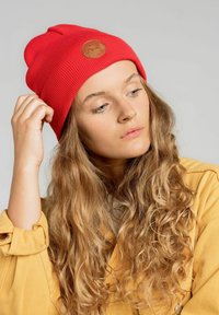 Czerwony, prążkowany beanie z okrągłą skórzaną łatą, noszony przez osobę z falującymi włosami, zestawiony z żółtą kurtką z kołnierzykiem.